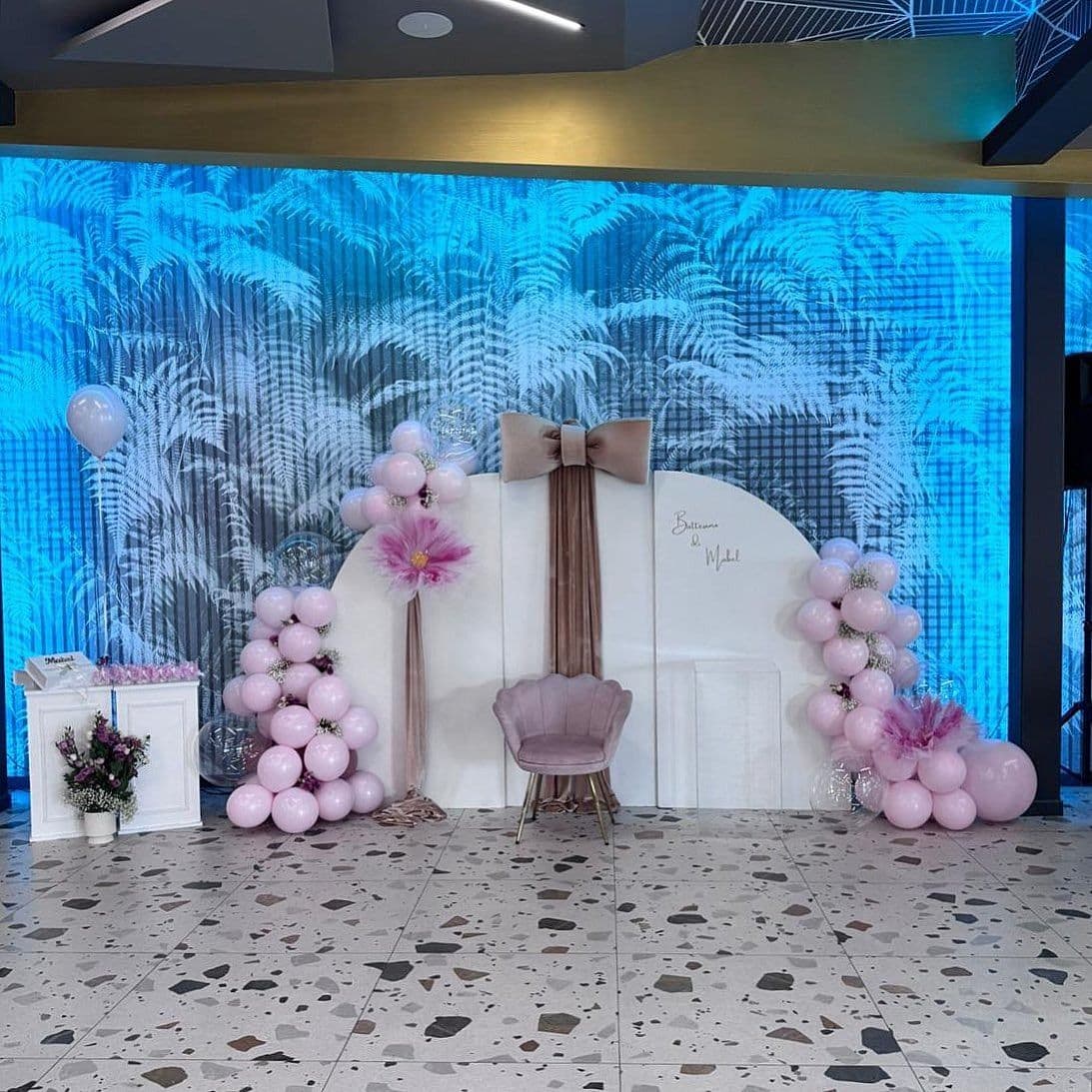 Allestimento in sala con fondale bianco, colonne di palloncini rosa e poltrona